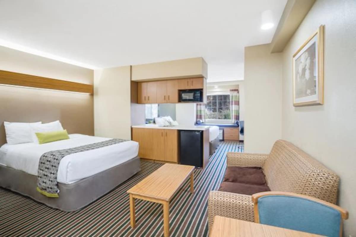 A compact hotel room with a bed, a small kitchenette, a seating area, and a window.