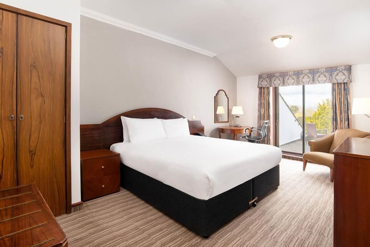 A hotel room with a large bed, a wooden wardrobe, a desk, a chair, and a balcony with a view.