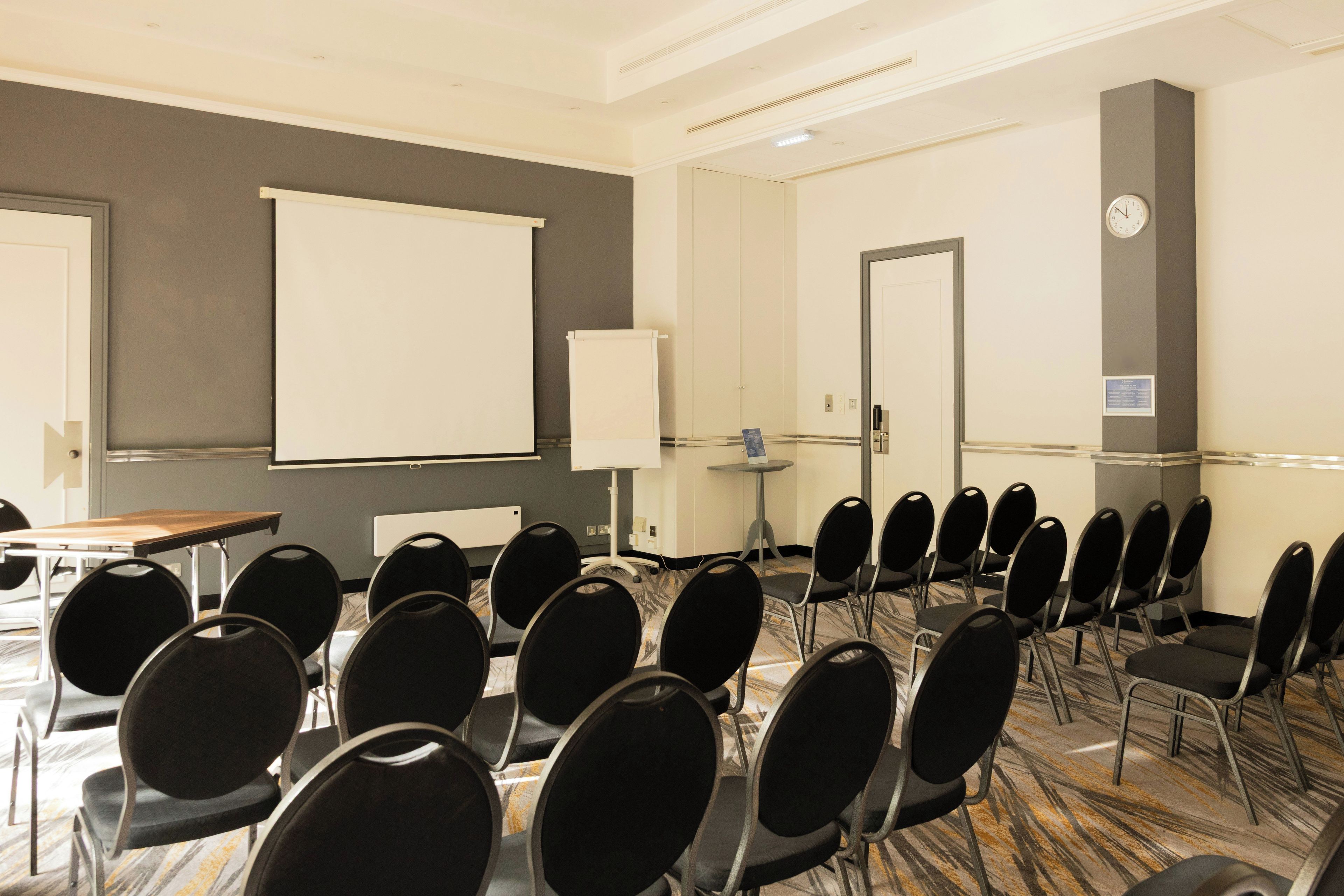 Un salón de conferencias con hileras de sillas, una pantalla de proyector y un reloj en la pared.