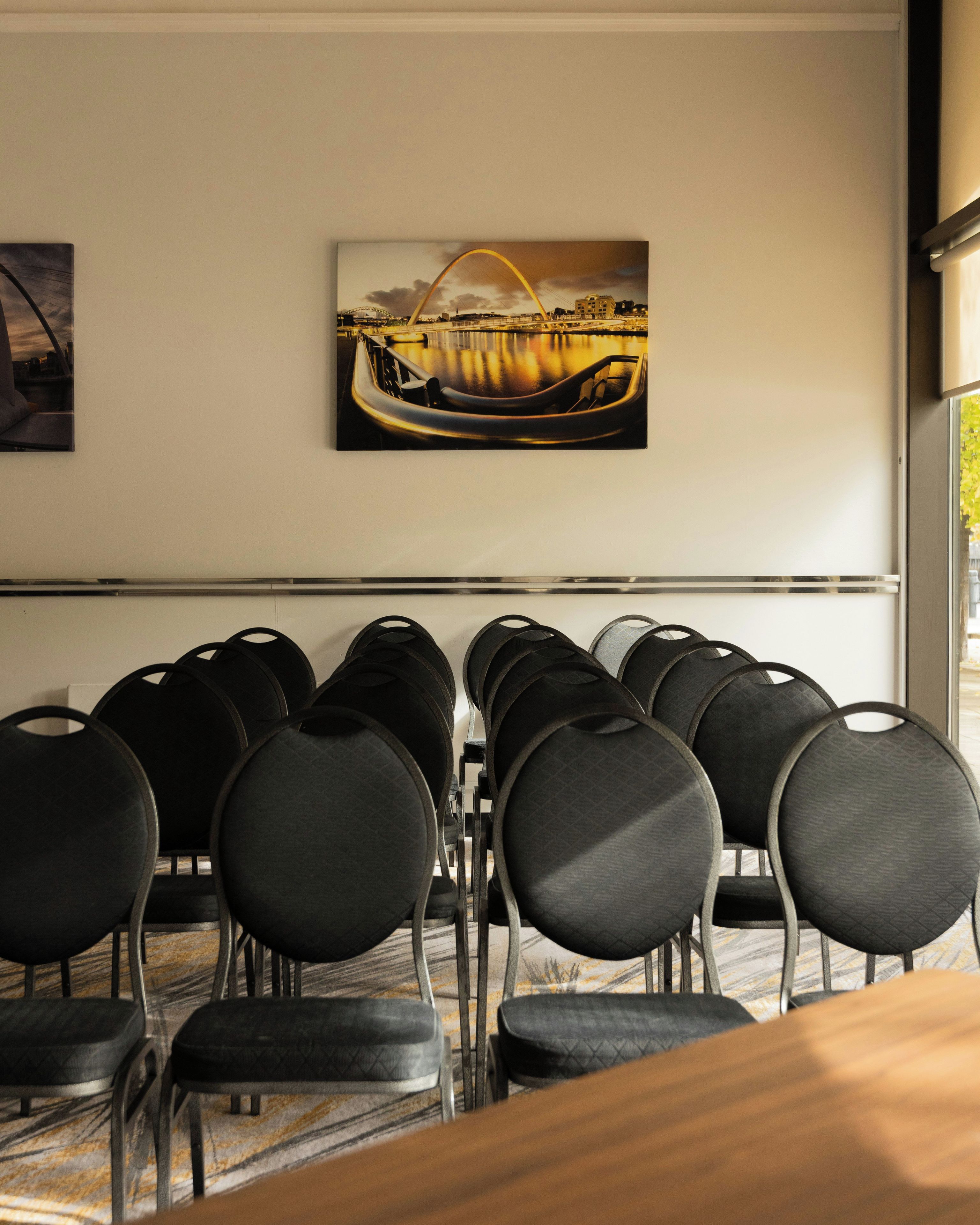 Une salle de conférence avec des rangées de chaises noires, un grand tableau représentant un pont et une fenêtre donnant sur des arbres.