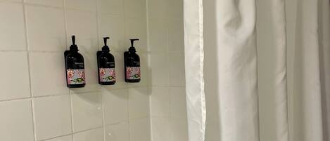 Un baño con una bañera blanca, un cabezal de ducha manual y tres botellas de champú en la pared.
