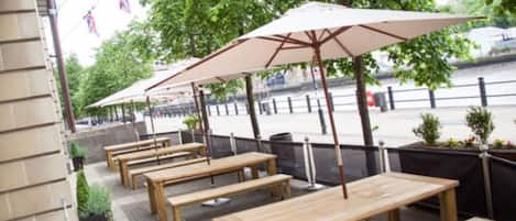 Outdoor seating with wooden tables and umbrellas, a paved walkway, and a river in the background.