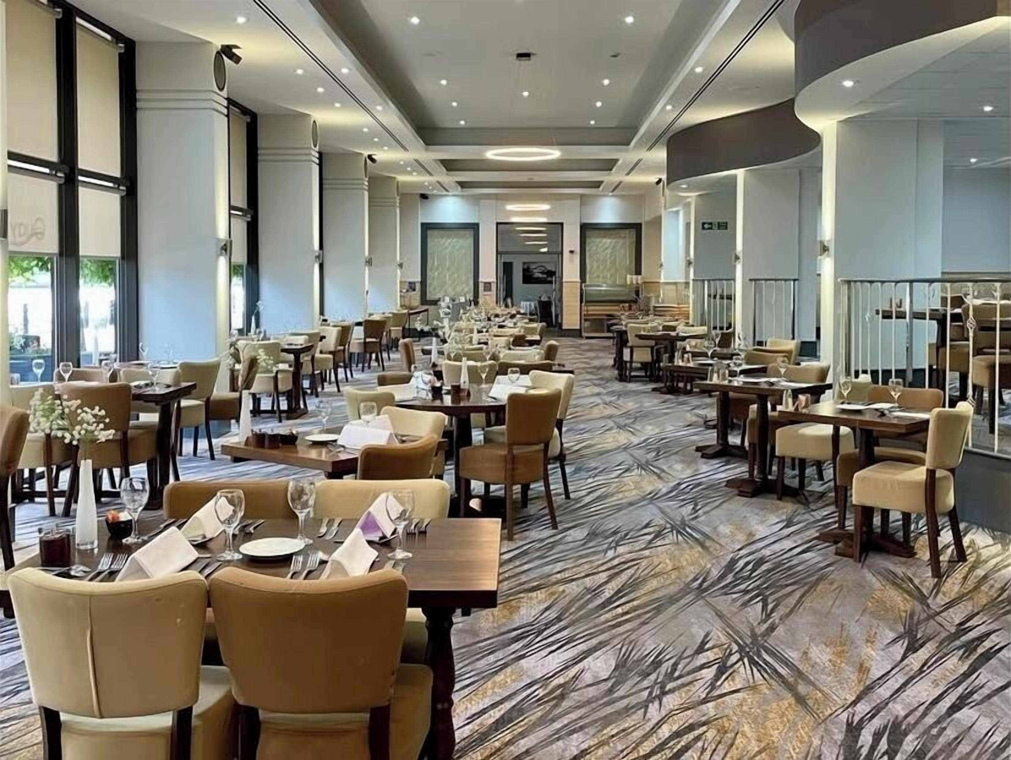 A spacious dining area with multiple tables and chairs, a patterned carpet, and large windows allowing natural light.