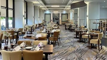 A spacious dining area with multiple tables and chairs, a patterned carpet, and large windows allowing natural light.