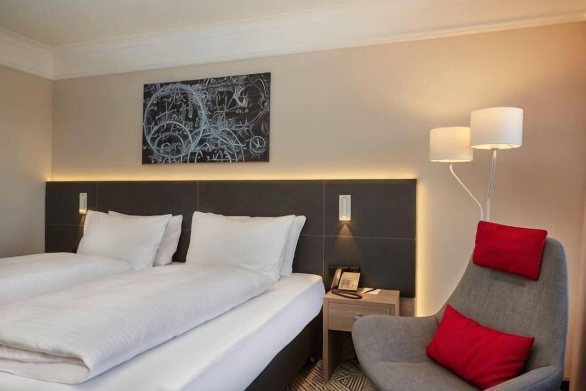 A hotel room with a large bed, a desk with a lamp, and a chair with red cushions.