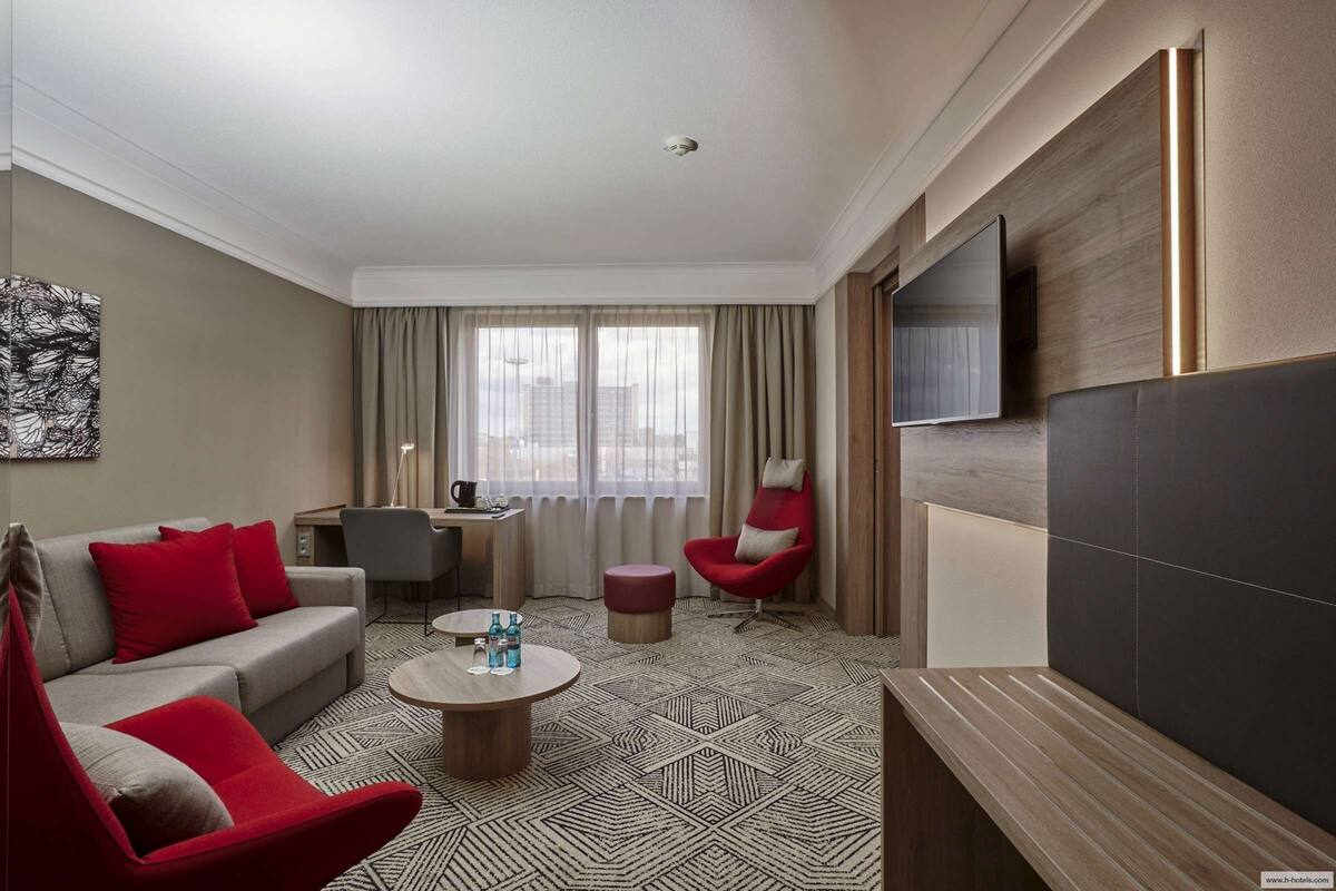 A modern hotel room with a sofa, red armchair, and a desk with a chair. There is a TV mounted on the wall and a window with curtains.