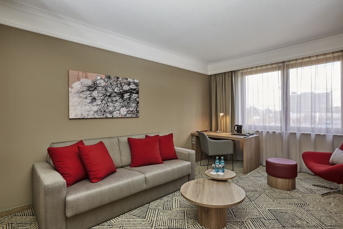 A modern hotel room with a sofa, red cushions, a round coffee table, a desk with a chair, and a large window with curtains.