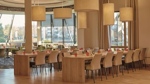 A spacious dining area with multiple tables and chairs, large windows, and hanging pendant lights. - H4 Hotel Hannover Messe (Laatzen)