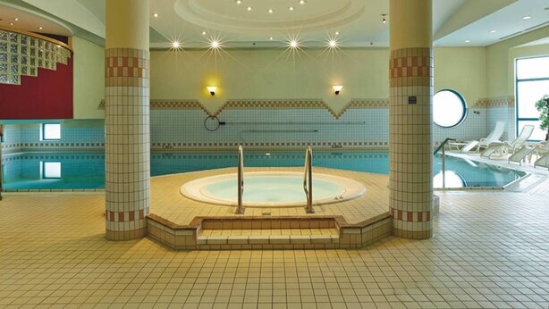 A large indoor swimming pool with a hot tub in the center, surrounded by tiled walls and columns. - H4 Hotel Hannover Messe (Laatzen)