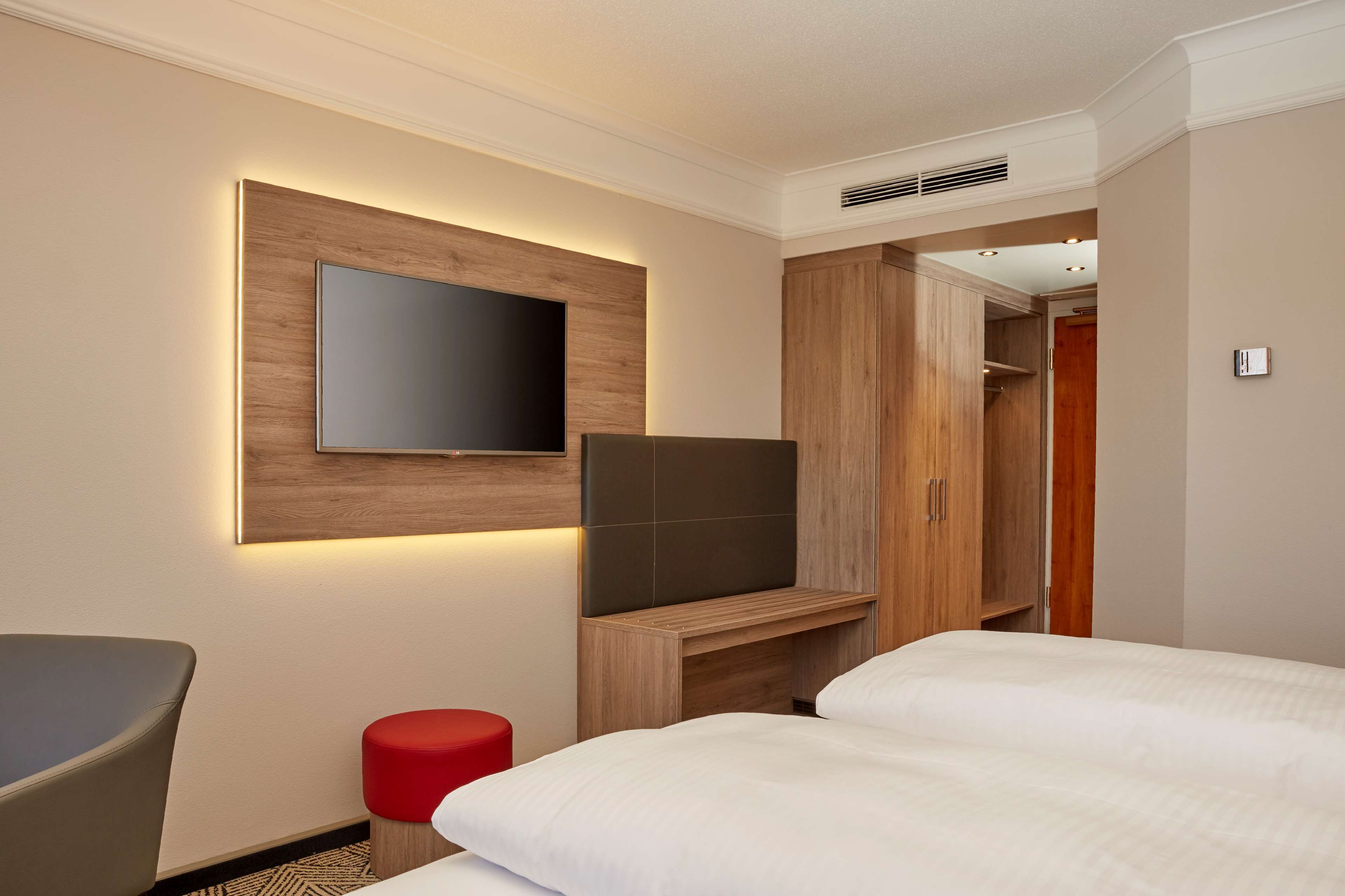 Una habitación de hotel con una cama, un televisor instalado en un panel de madera, un taburete rojo y un armario.