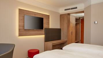 Una habitación de hotel con una cama, un televisor instalado en un panel de madera, un taburete rojo y un armario.
