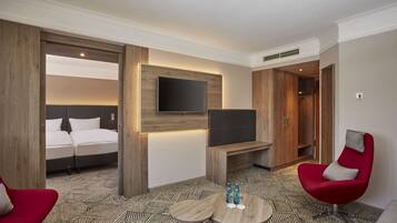 A hotel room with a bed, a TV mounted on the wall, a red armchair, a small round table with bottles, and a patterned floor.