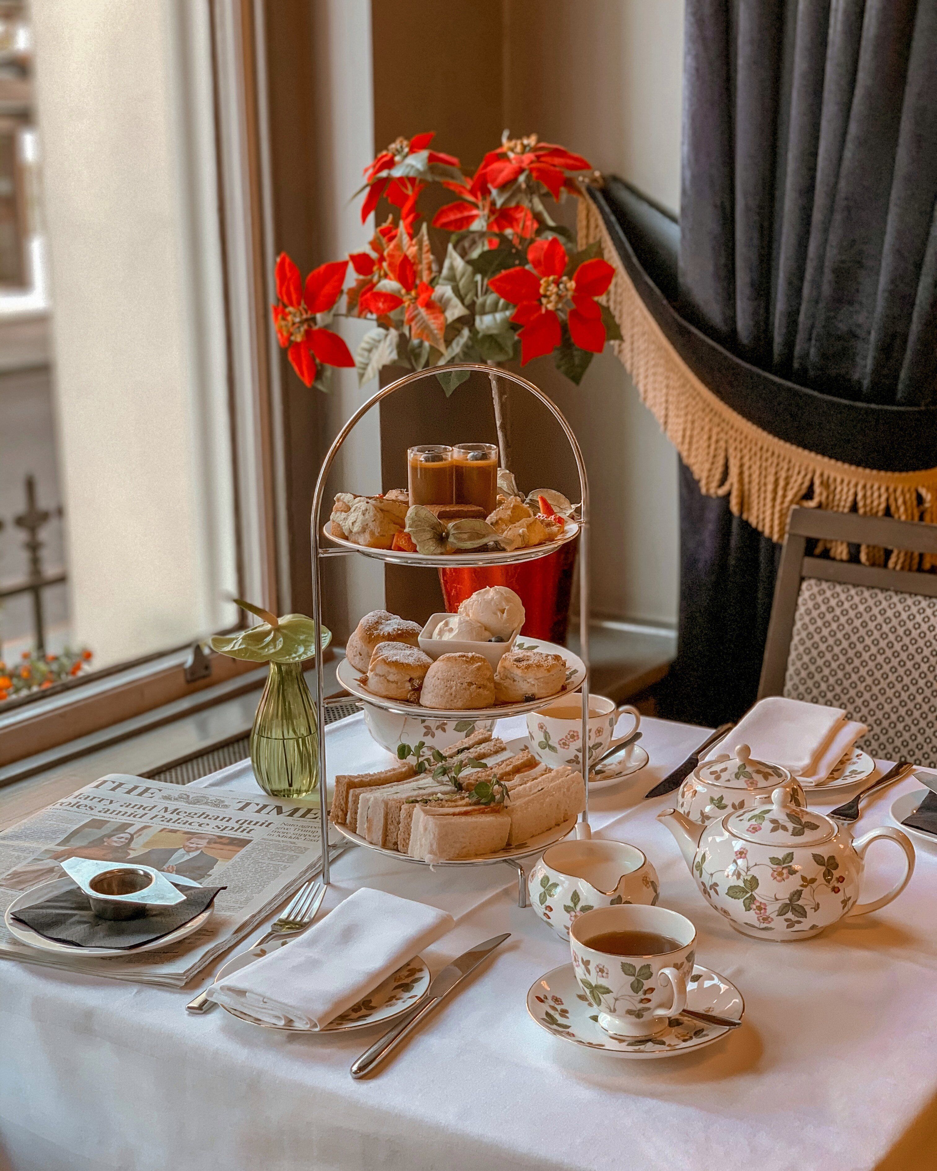 Een tafel gedekt voor afternoon thee, met een etagère vol gebak, een bloemenpot theepot en een vaas rode bloemen.