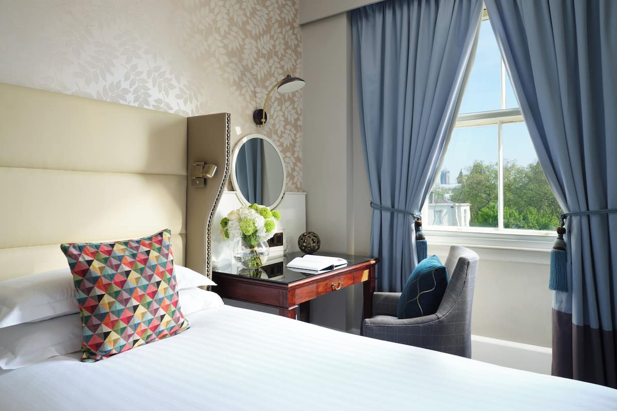 Habitación de hotel con cama, escritorio, silla, espejo y ventana con cortinas.