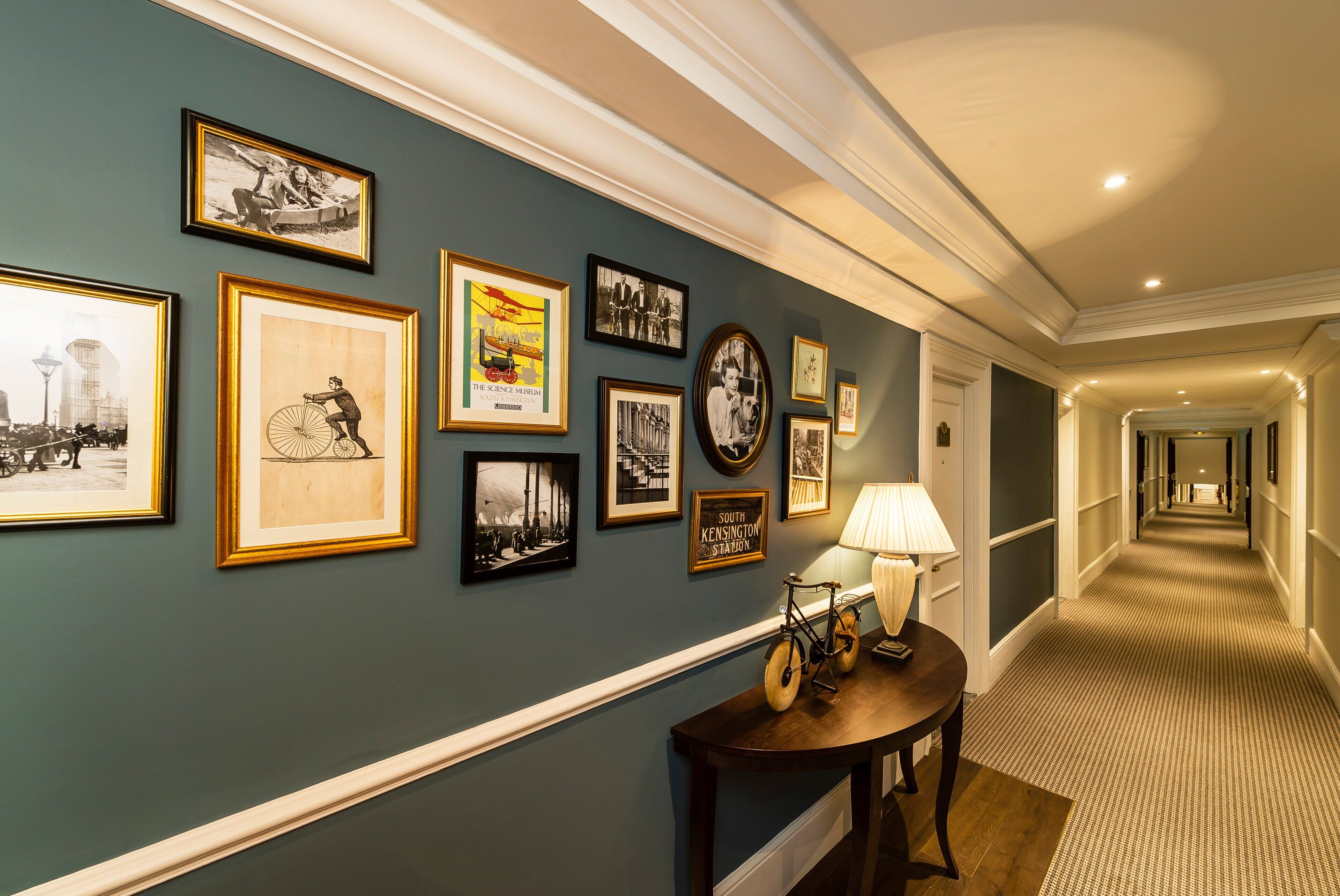 A hallway with framed pictures on the wall, a lamp on a table, and a carpeted floor.