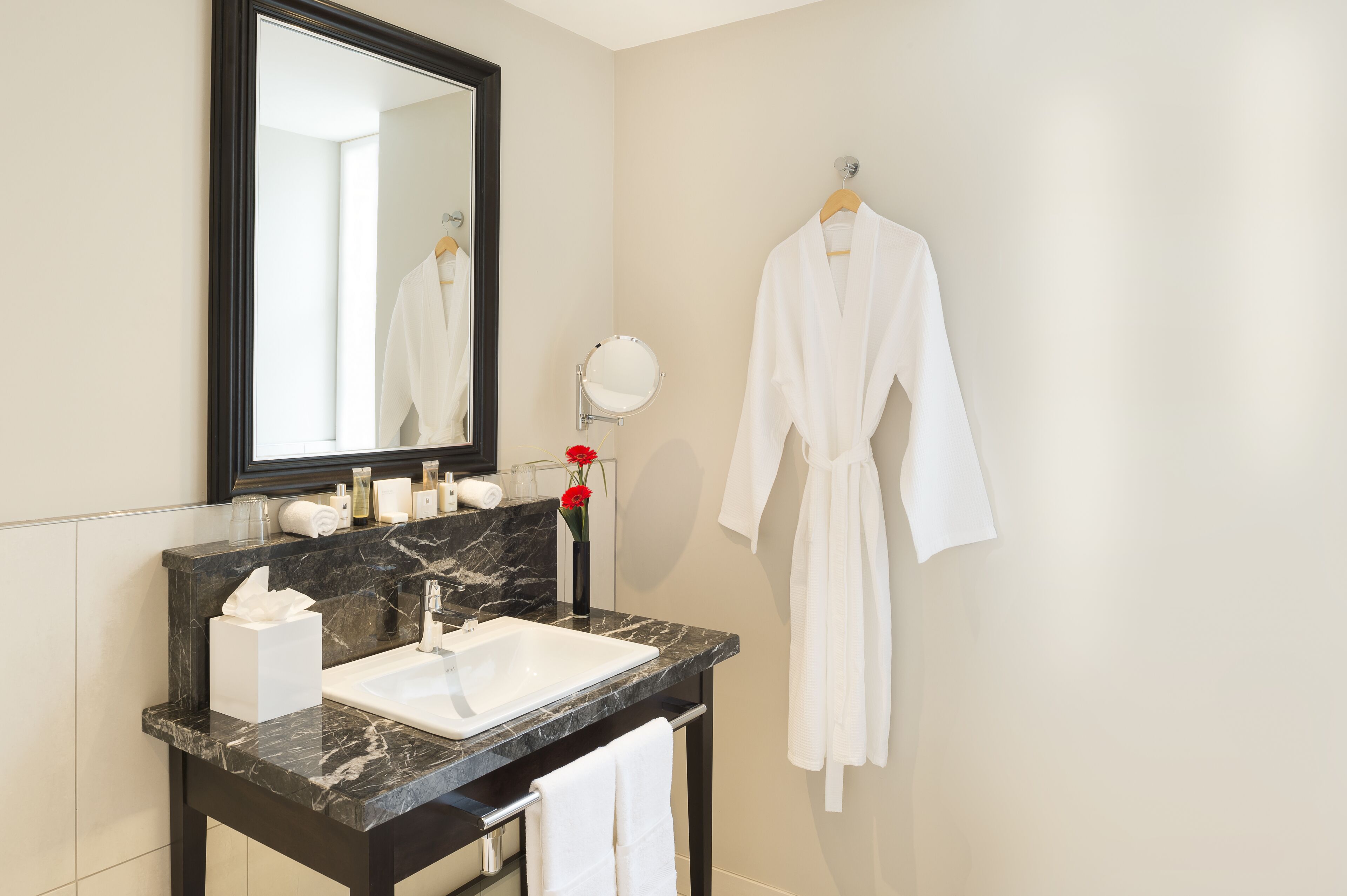 A bathroom with a marble sink, a mirror, a robe hanging on the wall, and a vase with red flowers.