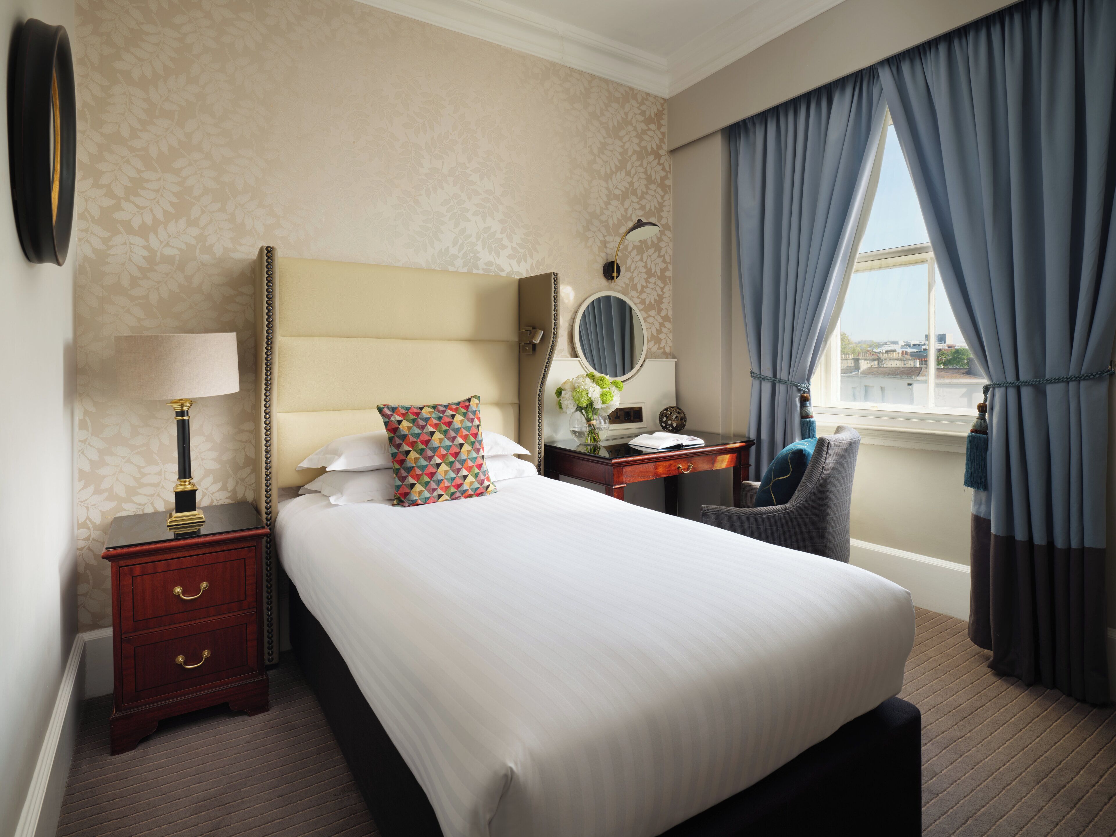Une chambre d’hôtel avec un grand lit, une table de chevet avec une lampe, une chaise, un bureau avec un miroir et une vue sur la ville par la fenêtre.