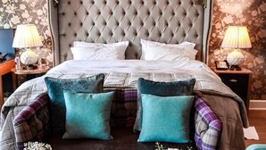 A bedroom with a tufted headboard, a bed with white linens, teal and plaid pillows, a bedside table with a lamp, and a breakfast setup on a brown ottoman.