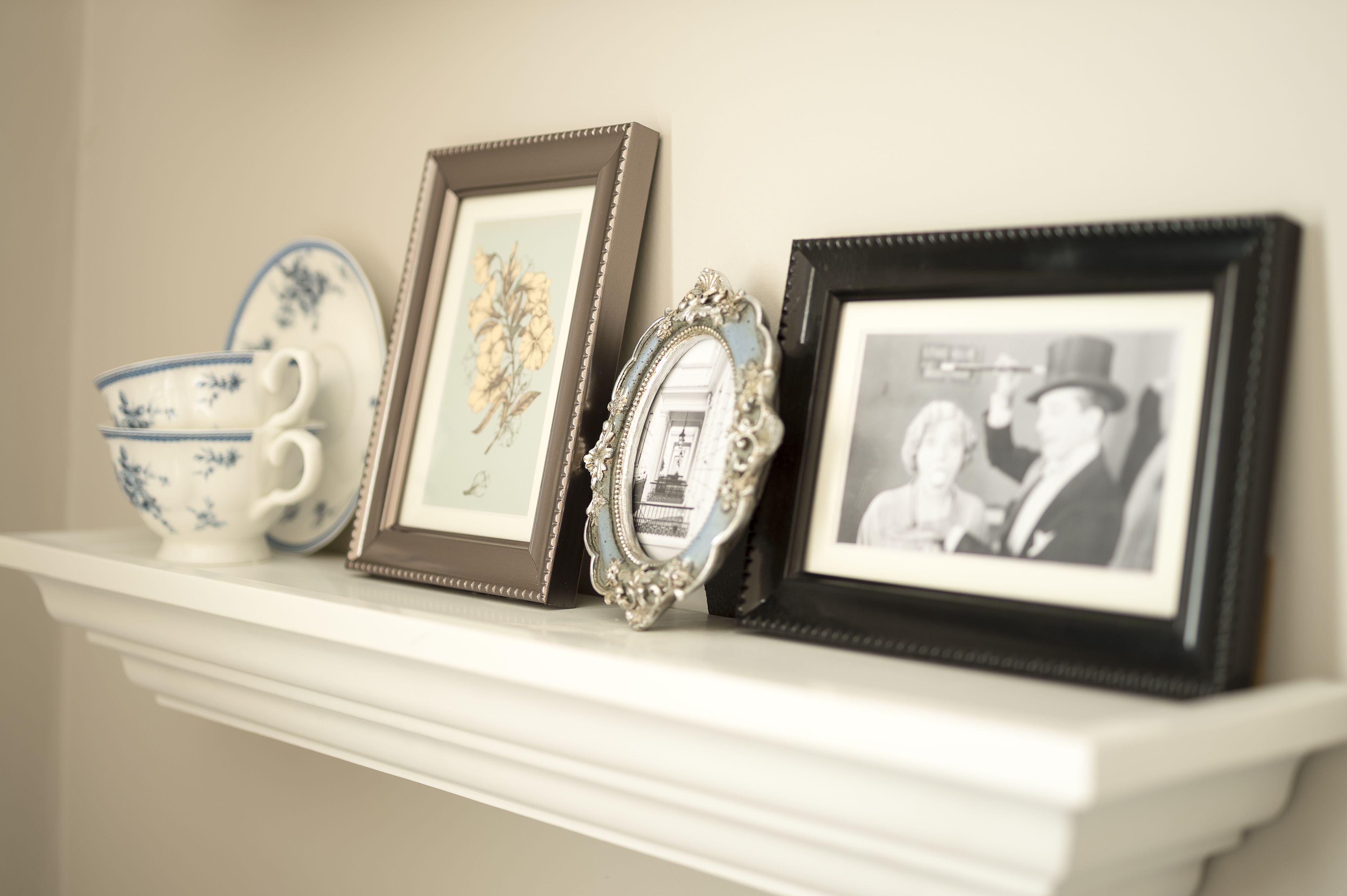A white mantelpiece with three framed items: a floral print, a mirror, and a black-and-white photo.