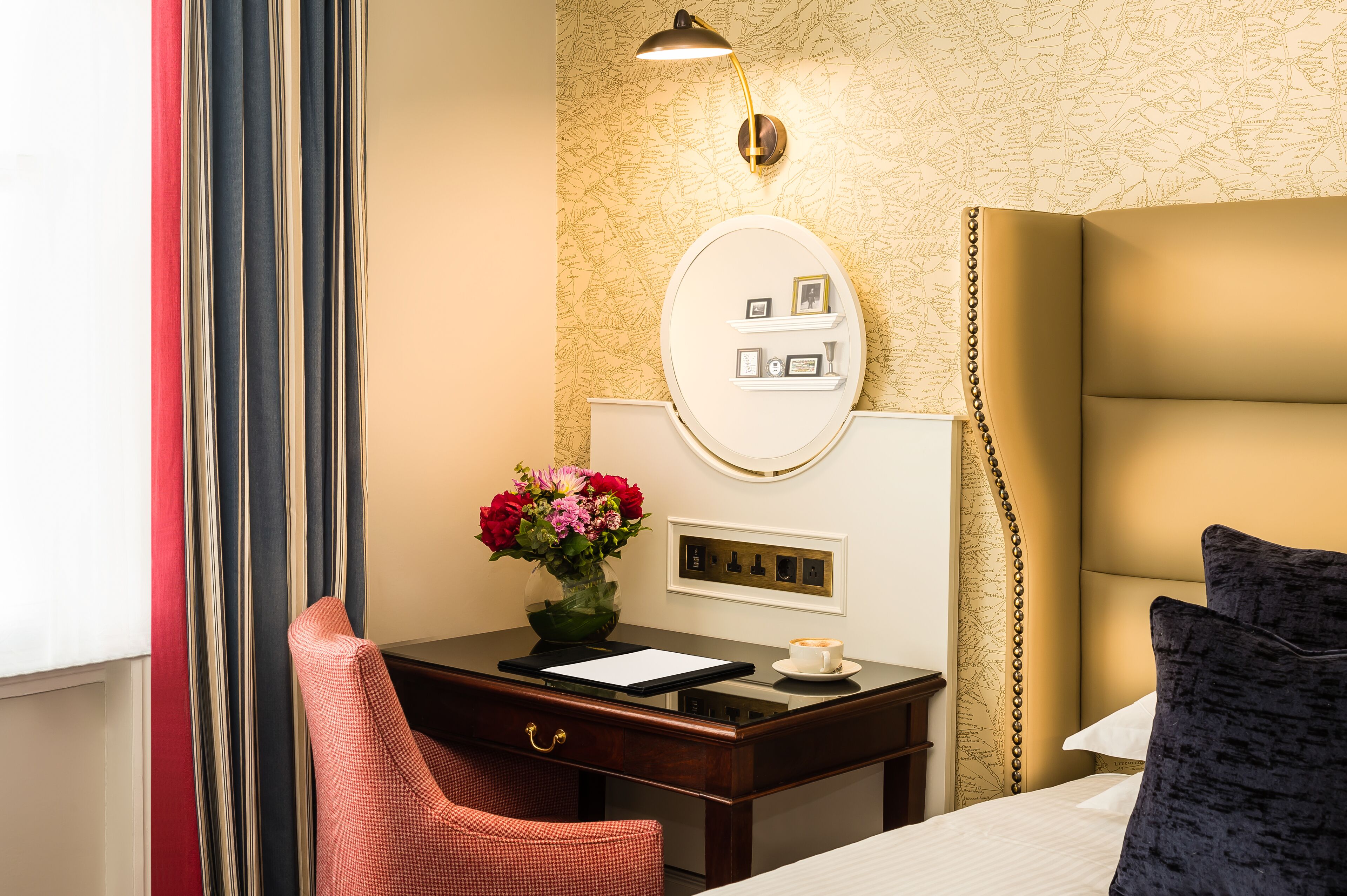 Een hotelkamer met een bed, een bureau met lamp, een stoel, een vaas met bloemen en een wandplank met foto's.