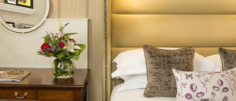 A hotel room with a wooden desk, a vase of flowers, a framed picture, a bed with white linens and decorative pillows.