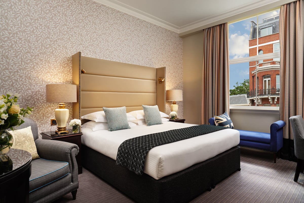 Una habitación de hotel con una cama grande, dos mesitas de noche con lámparas, una silla, un sofá y vistas a un edificio a través de una ventana.