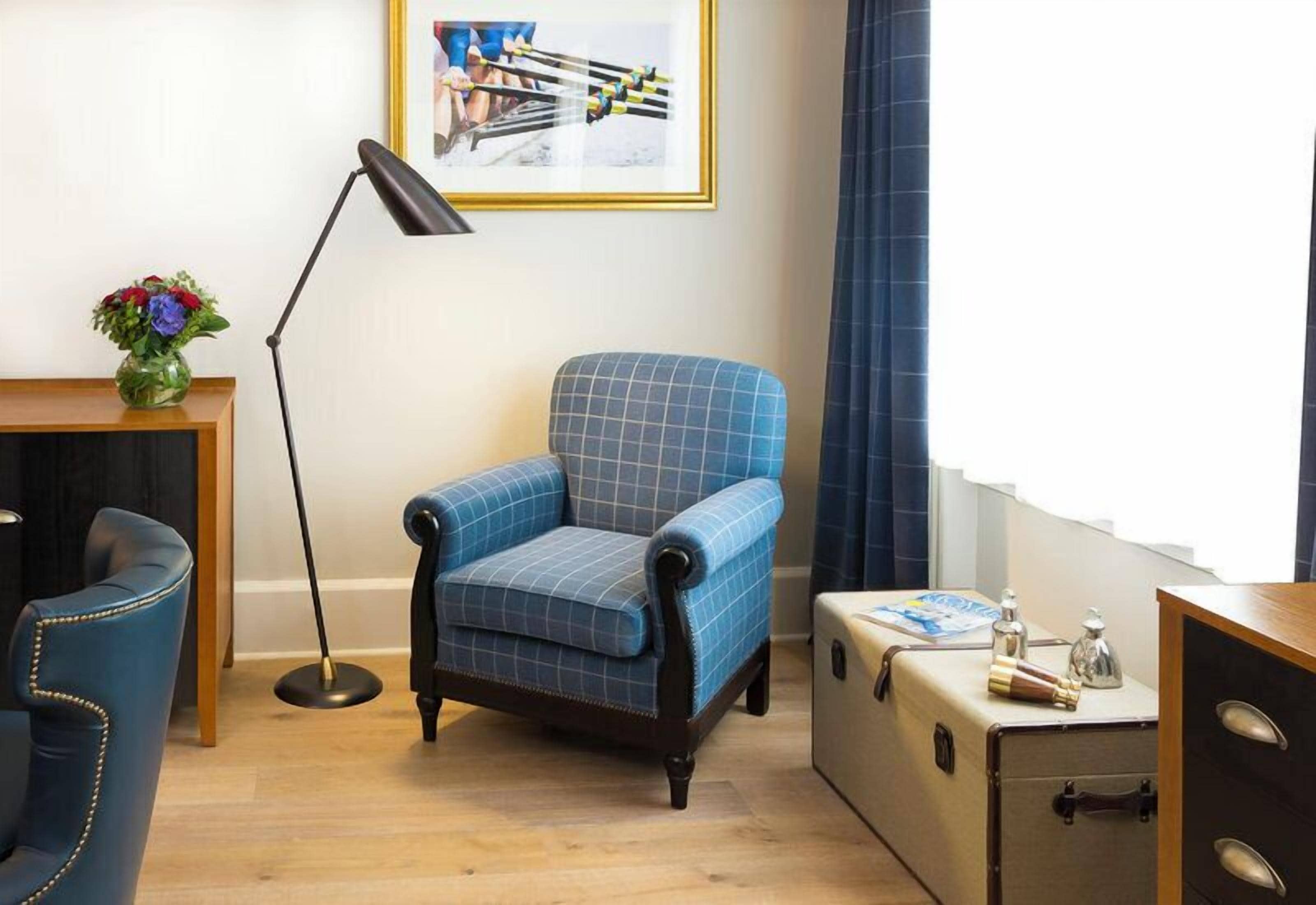 A room with a blue armchair, a floor lamp, a suitcase, and a framed picture on the wall.