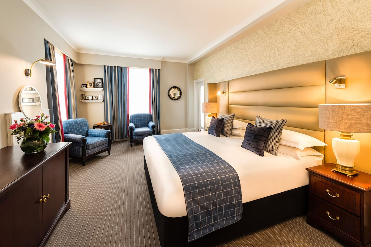 Una habitación de hotel con una cama grande, dos sillones, un aparador y una mesita de noche con una lámpara.