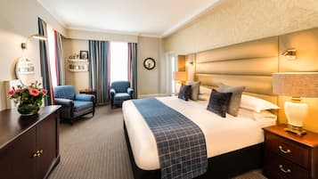 Una habitación de hotel con una cama grande, dos sillones, un aparador y una mesita de noche con una lámpara.