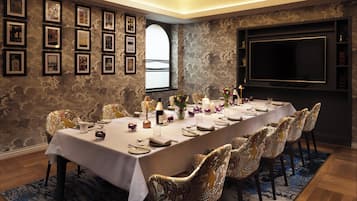 A long dining table set for a meal, surrounded by patterned chairs, with framed pictures on the walls and a large TV screen.