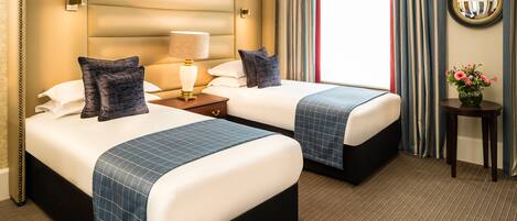 A hotel room with two beds, a nightstand with a lamp, a small table with a vase of flowers, and a large mirror on the wall.