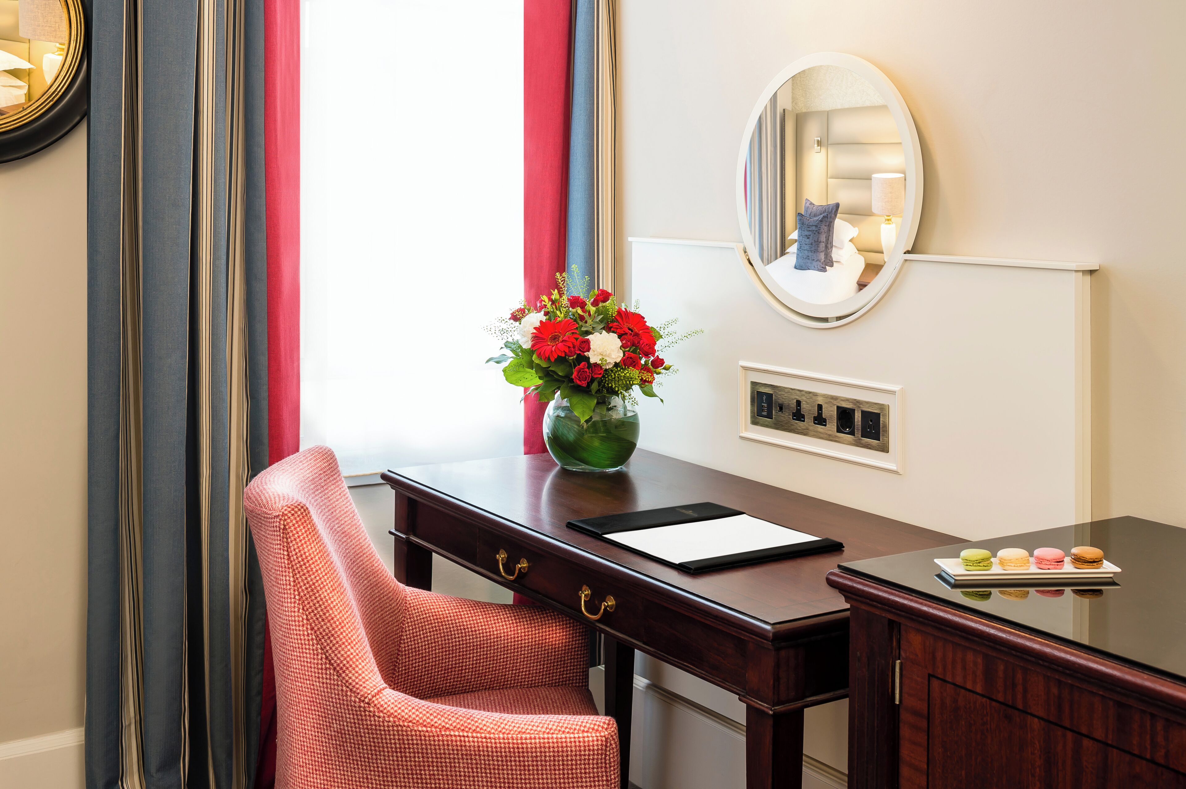 A hotel room with a desk, chair, mirror, vase with flowers, and a plate of macarons.