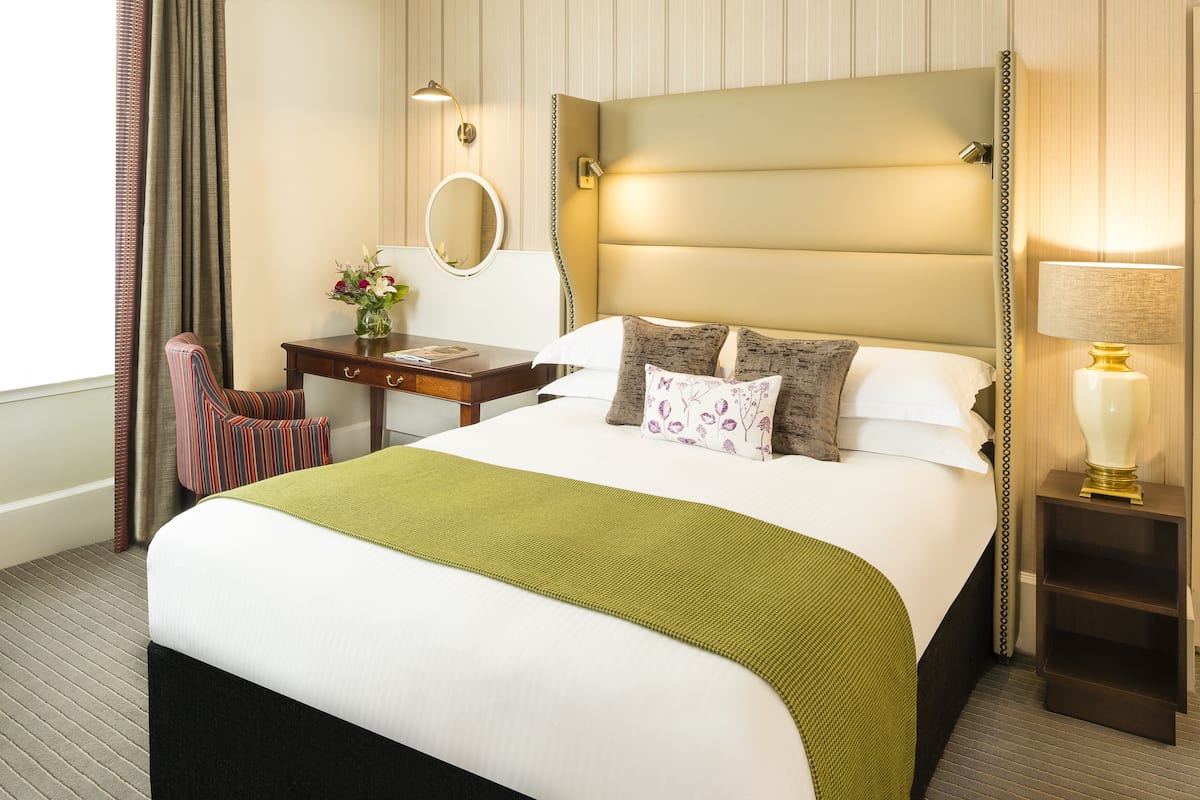 Habitación de hotel con una cama grande, un escritorio con silla, una mesita de noche con lámpara y un espejo en la pared.