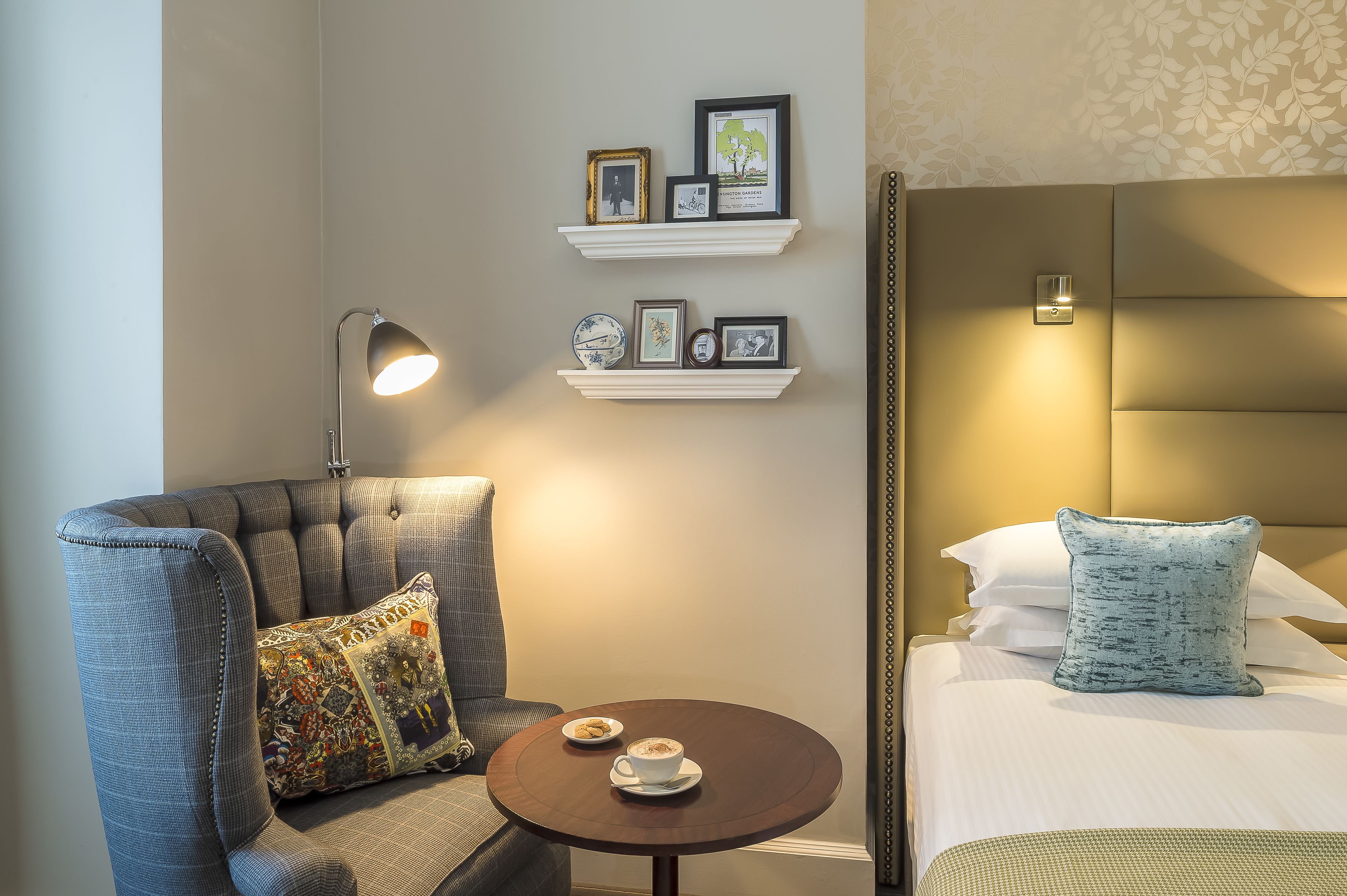 A hotel room with a tufted headboard, a round table with a cup and saucer, a sofa with a patterned cushion, and wall-mounted shelves with framed pictures.