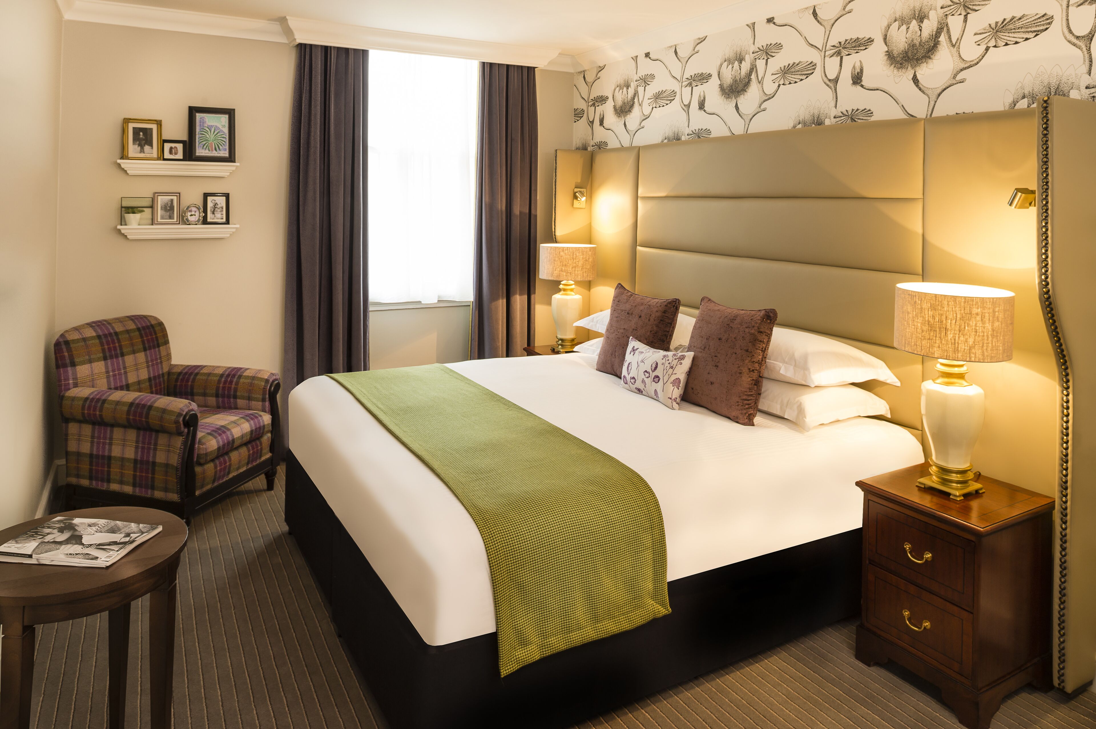 A hotel room with a large bed, two bedside lamps, a plaid armchair, a small table with a magazine, and a decorative headboard.