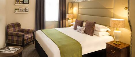 A hotel room with a large bed, two bedside lamps, a plaid armchair, a small table with a magazine, and a decorative headboard.