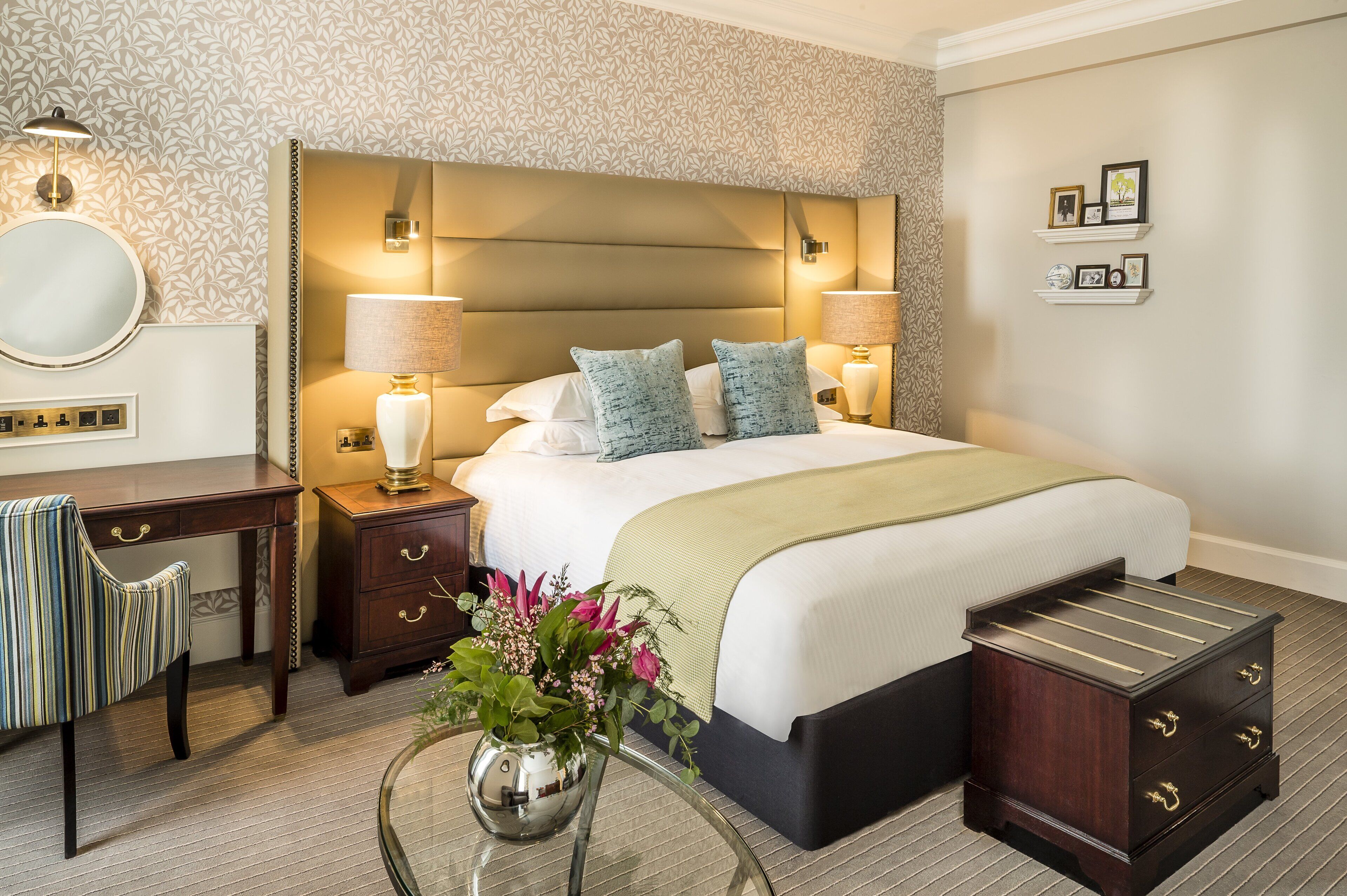A hotel room with a large bed, bedside lamps, a desk, and a small side table with a vase of flowers.