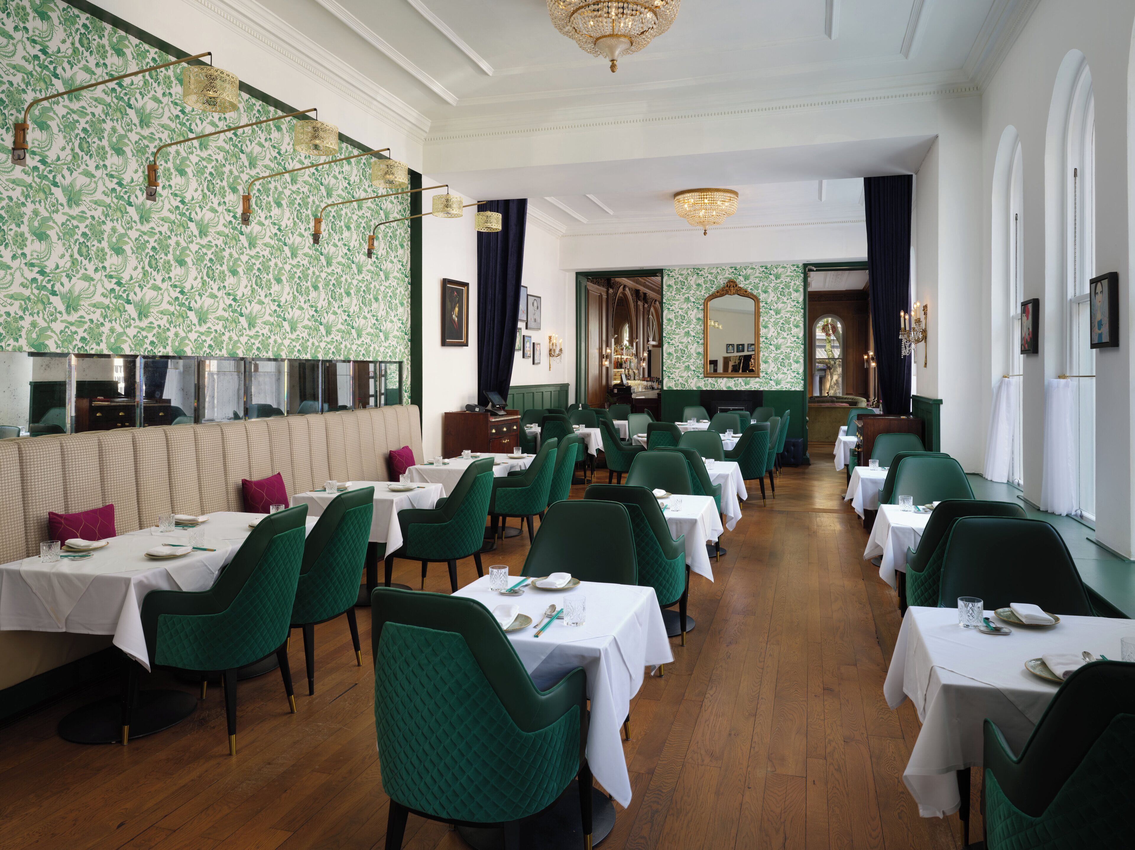 Restaurante com cadeiras estofadas em verde, toalhas de mesa brancas e papel de parede com padrão.