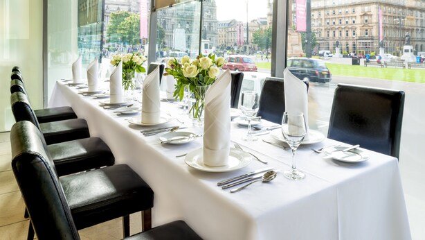 A restaurant with a long table set for a meal, chairs, and a view of the city through large windows.