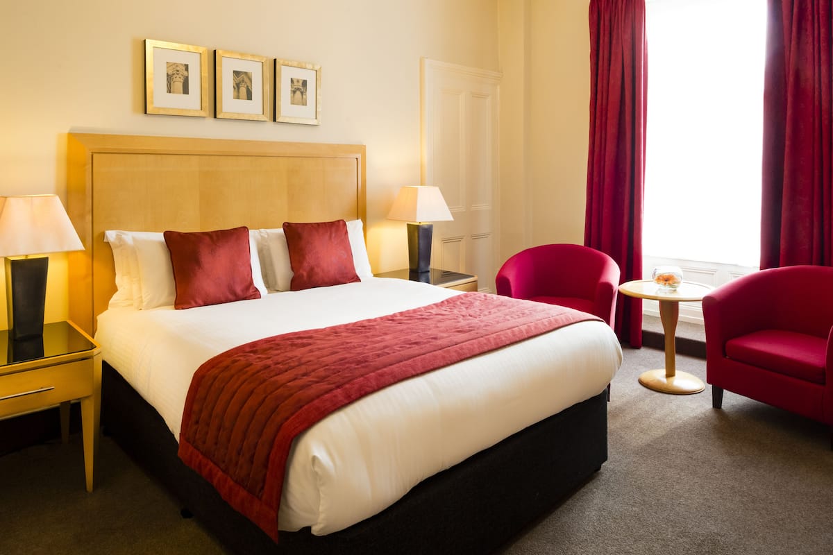 A hotel room with a large bed, two bedside tables with lamps, a red armchair, a small round table with a vase, and a window with red curtains.