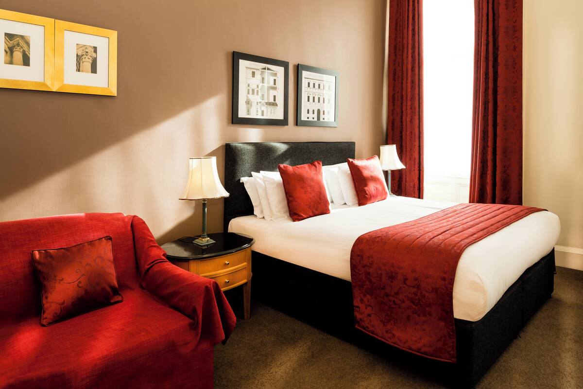 A hotel room with a bed, a nightstand, a red armchair, and two framed pictures on the wall.