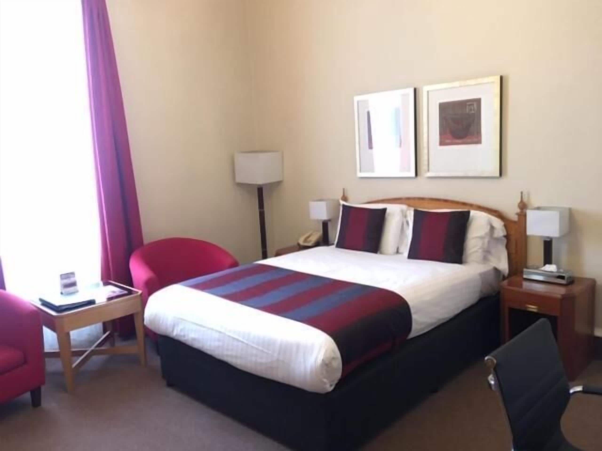 A hotel room with a bed, a desk, a chair, a lamp, and two framed pictures on the wall.