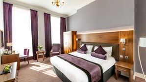 A hotel room with a large bed, a desk with a chair, a small table, and a lamp.