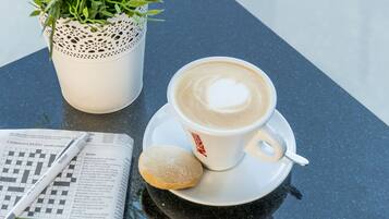 A cup of coffee with a heart design on the foam, a sugar cookie, a newspaper, and a potted plant on a table.