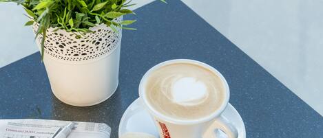 Une tasse de café sur laquelle on a dessiné un cœur dans la mousse, un biscuit sablé, un journal et une plante en pot, le tout posé sur une table.