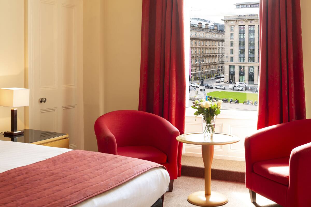 A hotel room with a bed, two red armchairs, a small table with a vase of flowers, and a view of a cityscape through large windows.