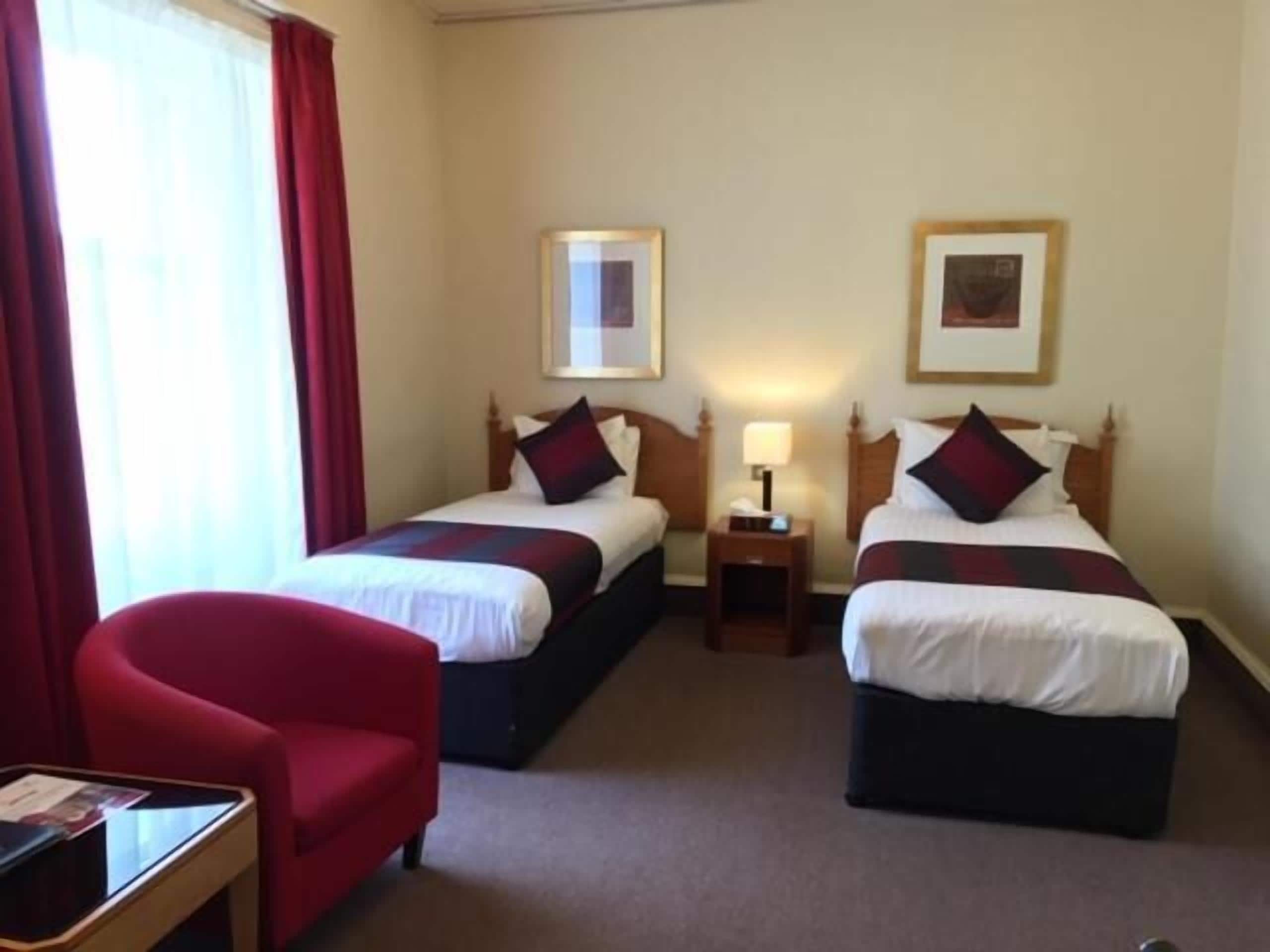 A hotel room with two single beds, a red armchair, a small table, and a nightstand with a lamp.