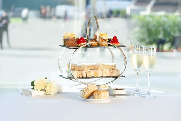 A tiered dessert stand with sandwiches and pastries, accompanied by two glasses of white wine, on a table with a view of a cityscape. - Millennium Hotel Glasgow (Glasgow)