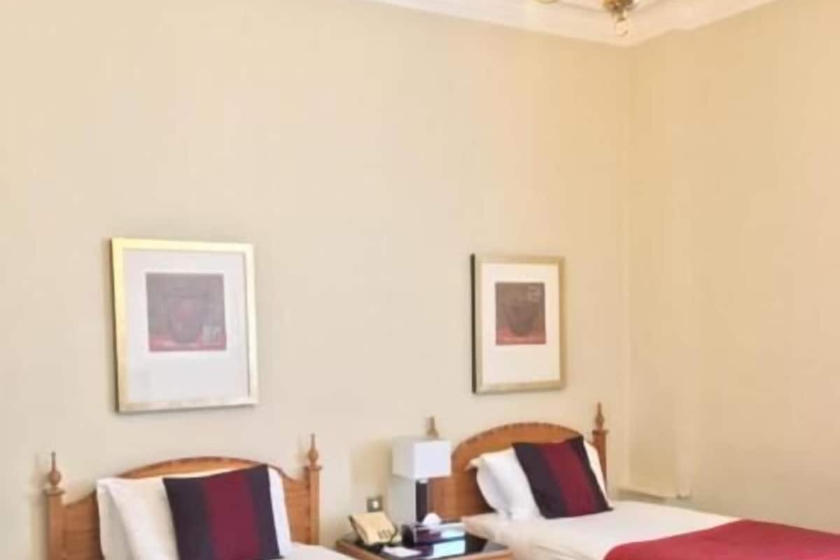 A hotel room with two beds, each with a red blanket, a wooden headboard, and a nightstand with a lamp. There is a chandelier on the ceiling and two framed pictures on the wall.