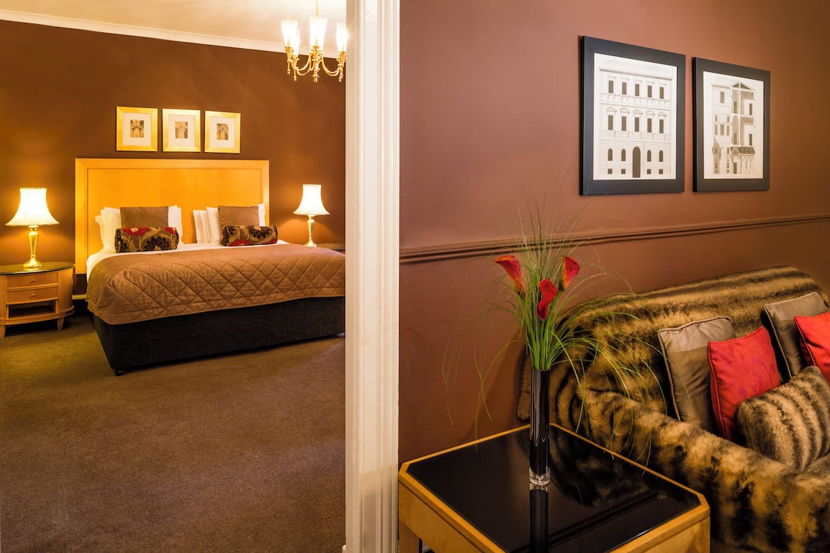 A hotel room with a bed, bedside lamps, a nightstand, a sofa, a vase with flowers, and two framed pictures on the wall.
